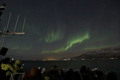 Soft green Aurora Borealis glowing above the ocean with Reykjavík city lights in the distance.